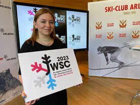 Eine junge Frau hält ein Schild mit dem Logo der Junioren-WM 2023 in St. Anton. Im Hintergrund sind historische Bilder des Ski-Club Arlberg zu sehen.