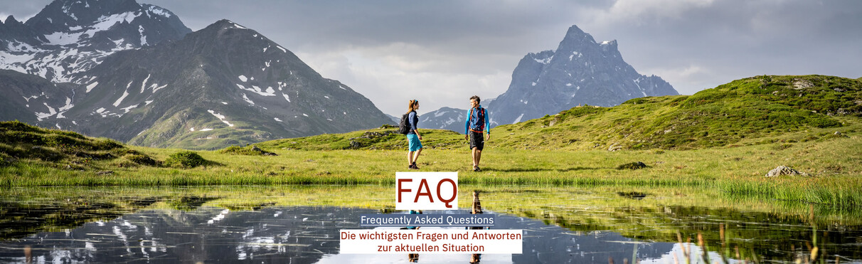 Zwei Wanderer stehen vor einem klaren Bergsee mit Blick auf eindrucksvolle Gipfel. In der Bildmitte ist die Aufschrift „FAQ – Frequently Asked Questions. Die wichtigsten Fragen und Antworten zur aktuellen Situation“.