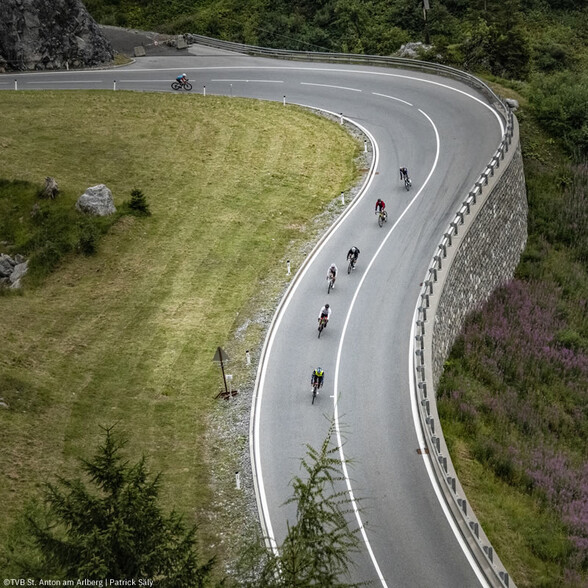 Mehrere Radfahrer fahren eine kurvige Serpentinenstraße durch eine alpine Landschaft hinab.