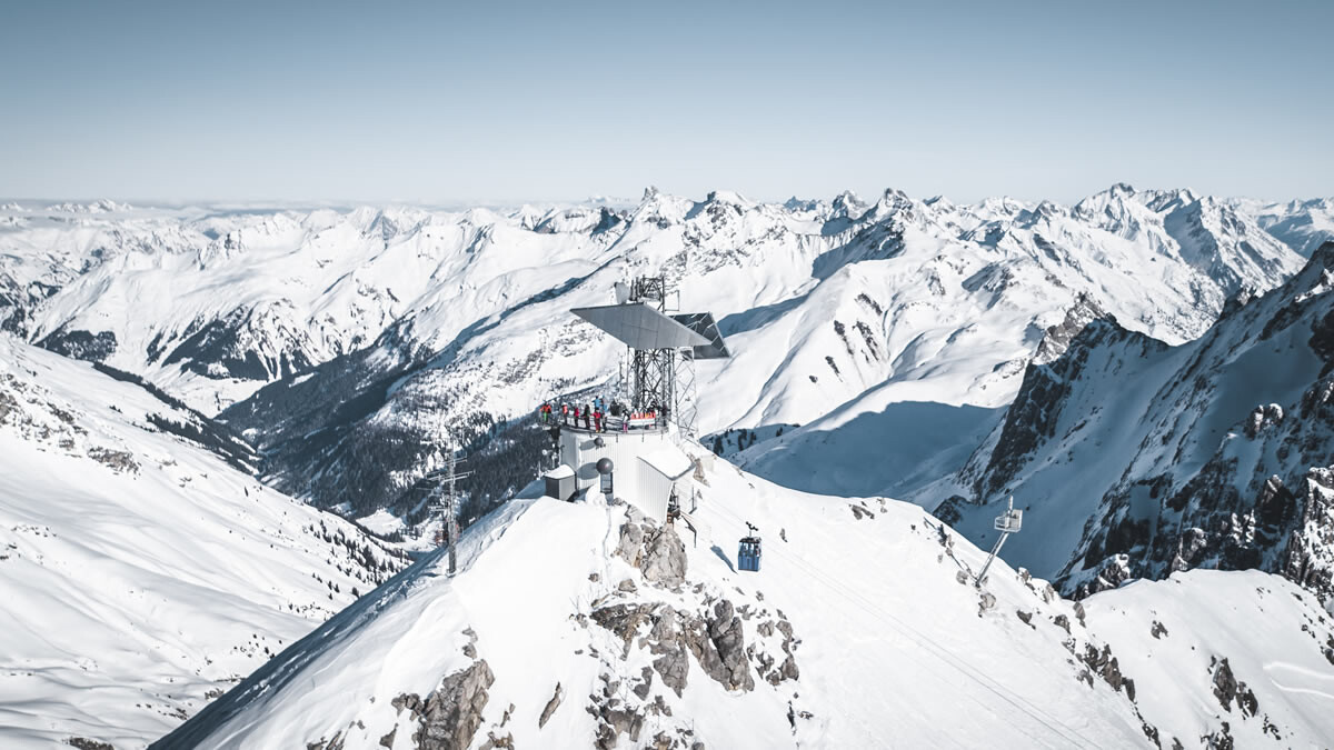 Person stands at Valluga platform in St. Anton, viewing a vast panorama of snowy mountain ranges.