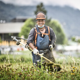 Ein Mann mit Schutzkleidung und Helm arbeitet mit einer Motorsense auf einer blühenden Wiese. Er lächelt in die Kamera.