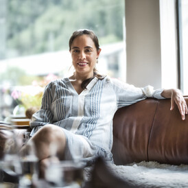 Eine Frau sitzt entspannt auf einem braunen Ledersofa in einem hellen Raum mit Ausblick auf grüne Berge.