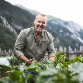 Ein lächelnder Mann steht in einem üppigen Gemüsegarten mit Bergkulisse. Er trägt ein olivgrünes Hemd und deutet mit der Hand auf das Gemüse.
