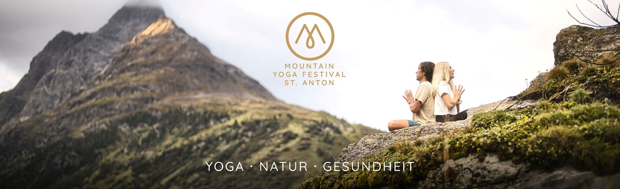 Two people meditating on a rock at the Mountain Yoga Festival in St. Anton, overlooking forested mountains.