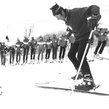 Schwarz-Weiß-Aufnahme von Edi Haueis in Aktion vor einer Reihe weiterer Skilehrer, alle tragen Uniformen mit Streifen auf der Jacke. Der Vordergrund zeigt den dynamischen Bewegungsstart auf Skiern.