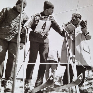 Schwarz-Weiß-Foto von Helmut Schranz mit Startnummer 4 am Starttor eines Skirennens, umgeben von zwei weiteren Personen. Alle tragen Winterkleidung aus den 1960er-Jahren.