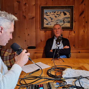 Max Kögl und Helmut Schranz nehmen eine Podcastfolge auf, einer spricht in ein Mikrofon, während der andere auf einem Holzstuhl in einer traditionell eingerichteten Stube sitzt. Im Hintergrund hängt ein Gemälde mit einer verschneiten Berglandschaft.