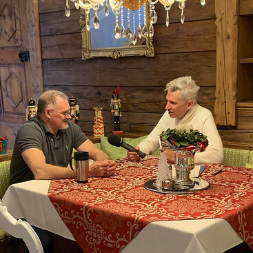 Christoph Schneider und Max Kögl sitzen an einem weihnachtlich geschmückten Holztisch mit rotem Tischläufer. Einer interviewt den anderen mit einem Mikrofon, im Hintergrund stehen Nussknackerfiguren.