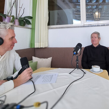 Max Kögl und  Edi Haueis sitzen an einem hell gedeckten Tisch in einem Raum mit großem Fenster. Ein älterer Herr mit schwarzem Pullover spricht in ein Mikrofon, auf dem Tisch steht eine Tasse Kaffee.