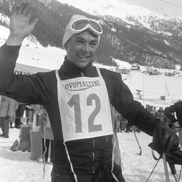 Ein junger Karl Schranz mit Startnummer 12 hebt lachend die Hand. Im Hintergrund sind schneebedeckte Berge und Zuschauende zu sehen.