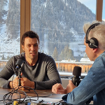 Mario Matt und Max Kögl führen ein Podcast-Interview an einem Tisch mit Blick auf verschneite Berge. Mikrofone und Tonaufnahmegeräte sind sichtbar.