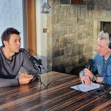 Mario Matt und Max Kögl unterhalten sich bei einem Podcast-Interview an einem Holztisch. Im Hintergrund ein Steinofen und warme Lichtstimmung.