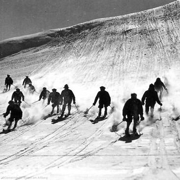 Skifahrer fahren in breiter Formation eine schneebedeckte Piste hinunter. Der Schnee wird aufgewirbelt, während die Gruppe sich dynamisch bewegt.