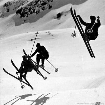 Drei Skifahrer in der Luft bei einer Synchronsprung-Darbietung. Ihre Positionen zeigen eine akrobatische Flugphase, während der Hintergrund verschneite Berghänge zeigt.