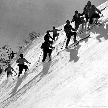 Eine Gruppe von Skifahrern in alter Skibekleidung erklimmt gemeinsam einen verschneiten Hang. Die Sonne wirft lange Schatten auf den Schnee.