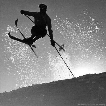 Ein Skifahrer springt dynamisch über einen Schneehang, während Schnee hinter ihm aufgewirbelt wird. Die Silhouette hebt sich gegen den Himmel ab.