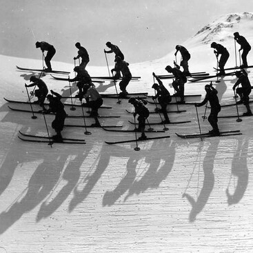 Eine große Gruppe Skifahrer startet synchron nebeneinander einen Abfahrtslauf. Ihre Bewegungen und Schatten auf dem Schnee ergeben ein starkes Muster.