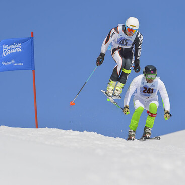Zwei Skifahrer:innen springen gleichzeitig über eine Pistenkante beim Skirennen „Der Weisse Rausch“. Links ist ein blauer Torflaggenpfosten mit der Aufschrift „Der Weisse Rausch“ zu sehen. Der Himmel ist wolkenlos blau.