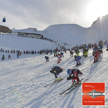 Zahlreiche Skifahrer:innen starten gleichzeitig beim Skirennen „Der Weisse Rausch“ in St. Anton am Arlberg. Sie fahren in Hocke eine steile, verschneite Piste hinunter, während Zuschauer:innen am Pistenrand zusehen. Die Sonne scheint über ein alpines Winterpanorama.
