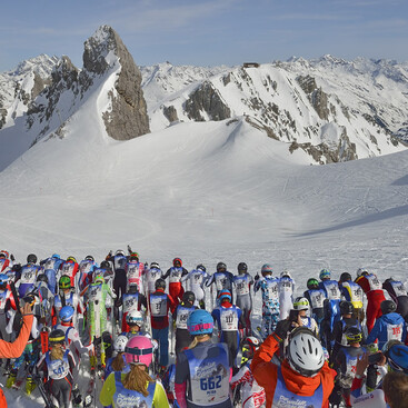 Eine große Gruppe Skifahrer:innen steht am Gipfel vor dem Start eines Skirennens in St. Anton am Arlberg. Im Hintergrund ragen schroffe, schneebedeckte Felsen und alpine Gipfel empor.