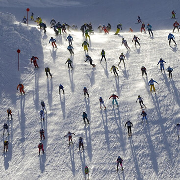 Skifahrer:innen beim Skirennen „Der Weisse Rausch“ starten gleichzeitig aus verschiedenen Richtungen auf einer breiten Piste. Das Bild zeigt die Szene aus der Vogelperspektive mit langen Schatten auf dem Schnee.