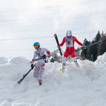 Zwei Skirennläufer:innen tragen ihre Skier über eine Schneekante bei einem Hindernis des Rennens „Der Weisse Rausch“ in St. Anton am Arlberg. Beide tragen Rennanzüge und Helme.