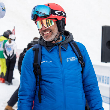 Peter Mall in blauer Winterjacke, rotem Helm und verspiegelter Skibrille lächelt bei einem Skirennen in St. Anton am Arlberg. Im Hintergrund sind weitere Rennteilnehmer:innen zu sehen.