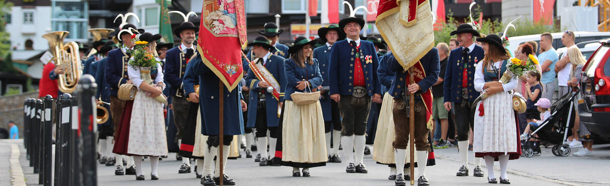 Musikkapelle St. Anton am Arlberg in traditioneller Tracht mit Fahnen steht auf einem Platz, im Hintergrund sitzen Zuschauer auf Bänken.