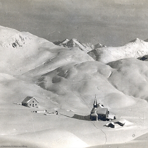 Eine historische Winteraufnahme zeigt eine kleine Kirche inmitten verschneiter Hügel in den Alpen.
