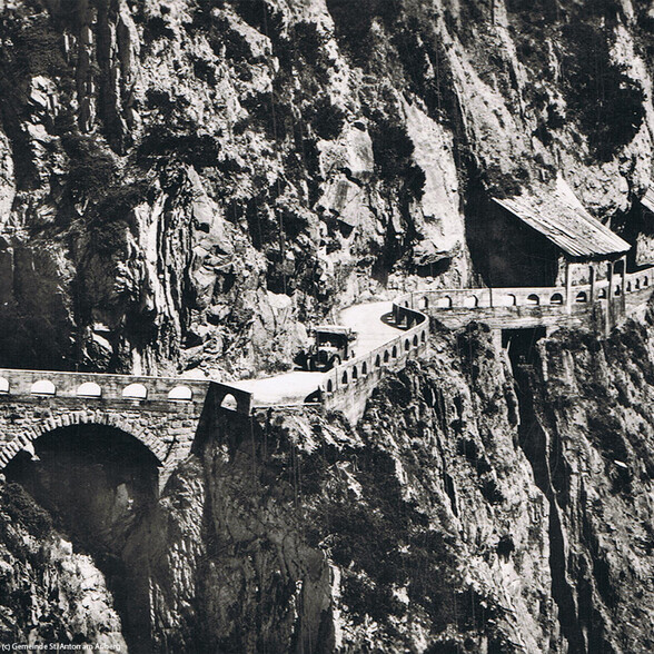 Ein altes Auto fährt auf einer gewundenen Straße mit Brücke und Steinschutzwand durch eine felsige Schlucht.