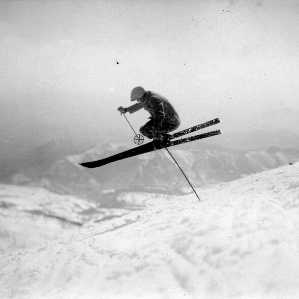 Schwarzweißaufnahme eines Skifahrers beim Sprung. Die Szene zeigt einen eleganten Telemark-Sprung vor verschneiten Bergen.