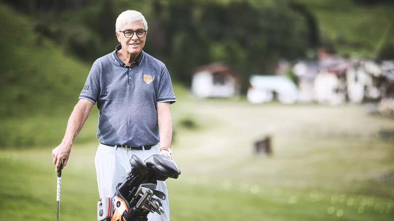 Ein älterer Herr lächelt in die Kamera und steht mit Golftasche und Schläger auf einem hügeligen Fairway. Der Hintergrund zeigt eine verschwommene Ortschaft in den Bergen.