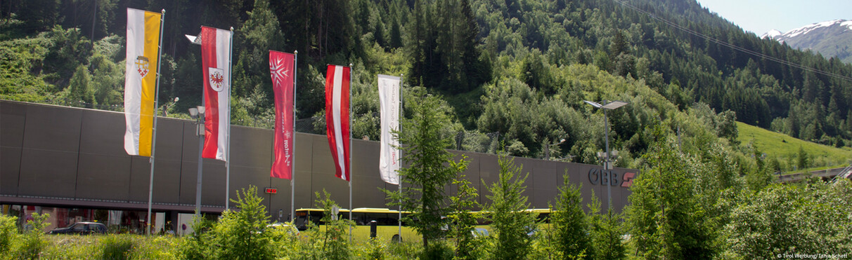Mehrere Fahnen vor dem modern gestalteten Bahnhof am Arlbergpass. Im Hintergrund dichte grüne Wälder und ein sommerlicher Alpenhang.