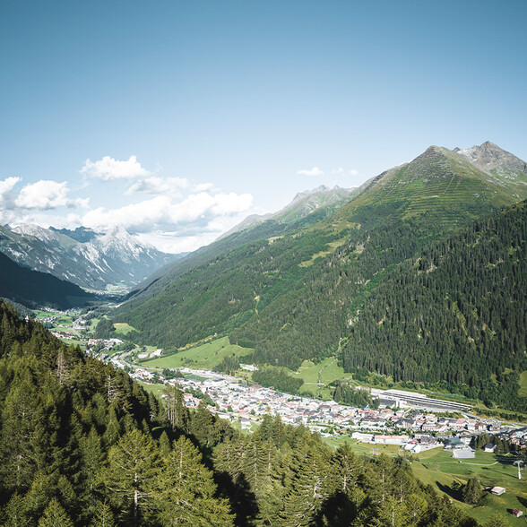 Panoramablick über das Stanzertal mit dem Ort St. Anton am Arlberg, umgeben von grünen Berghängen und hohen Gipfeln unter blauem Sommerhimmel.