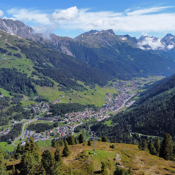 Atemberaubende Aussicht auf das Tal von St. Anton am Arlberg mit umliegenden Bergen, Wäldern und verstreuten Häusern, aufgenommen von einem Aussichtspunkt mit Gipfelkreuz.