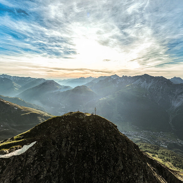 Die untergehende Sonne taucht den Himmel über einem alpinen Gipfel mit Kreuz in warmes Licht. Die umliegenden Berge zeichnen sich als dunkle Silhouetten ab.