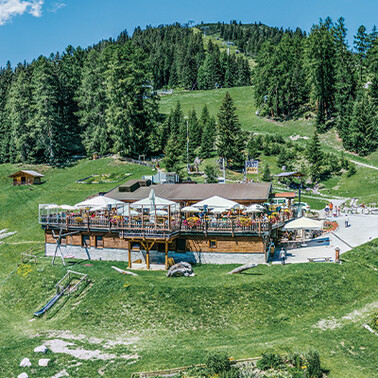 The rustic wooden alpine hut with a large terrace. It is located on a hillside in St. Anton am Arlberg, surrounded by green meadows and forests.