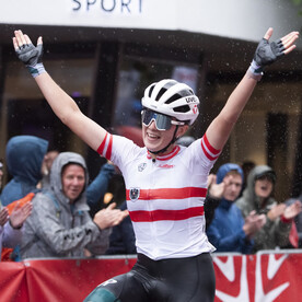Eine österreichische Radrennfahrerin jubelt mit erhobenen Armen beim Überqueren der Ziellinie.