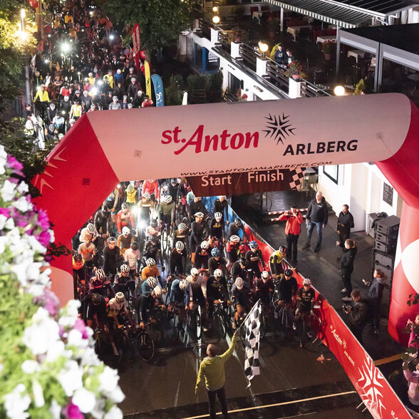 Der Start des Arlberg Giro unter einem großen roten Torbogen mit der Aufschrift „St. Anton am Arlberg“. Zahlreiche Radfahrer:innen warten auf das Startsignal.