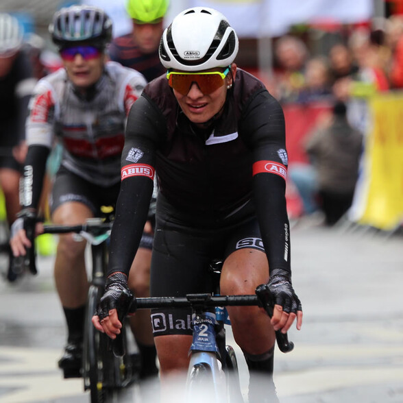 Eine Radfahrerin überquert die Ziellinie mit konzentriertem Blick, im Hintergrund jubelnde Zuschauer.
