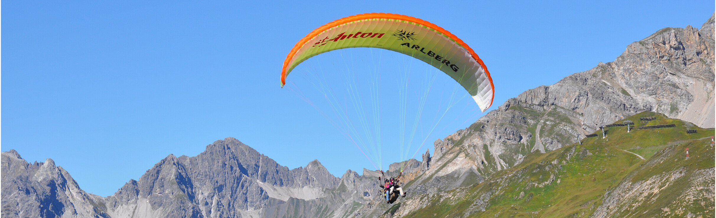 Gleitschirmflieger vor klarblauem Himmel und grüner Alpenlandschaft.
