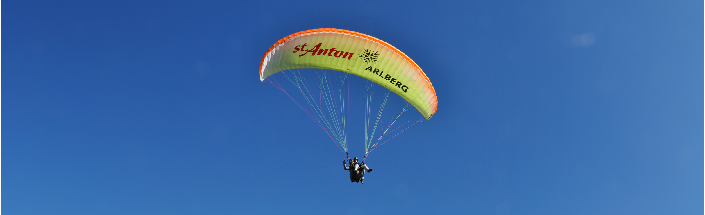 Gleitschirm mit orange-grünem Schirm vor wolkenlosem Himmel.