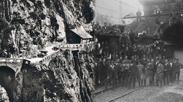 Historische Schwarz-Weiß-Aufnahme zeigt Arbeiter und alte Bahnanlagen beim Tunnelbau in steiler Felslandschaft.