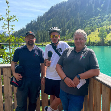 Alexander Klimmer, Max Kögl und Markus Strolz stehen lachend vor dem türkisblauen Verwallsee mit bewaldeten Berghängen im Hintergrund.