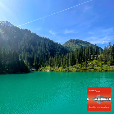 Der türkisgrüne Verwallsee liegt still unter einem strahlend blauen Himmel, umgeben von dichten Wäldern und grünen Bergen. Das Logo „Den Bergen lauschen“ befindet sich unten rechts.