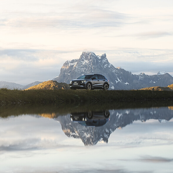 Ein schwarzer Audi steht vor imposanter Bergkulisse und spiegelt sich in einem klaren Bergsee. Die Szene vermittelt Ruhe und Kraft zugleich.