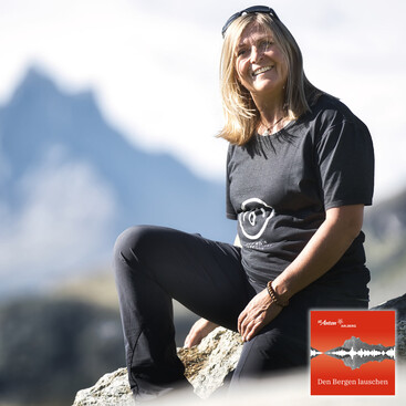 Geli Häusl sitzt lachend auf einem Felsen in alpiner Landschaft. Im Hintergrund sind verschwommene Berggipfel zu sehen. Sie trägt ein schwarzes T-Shirt mit einem stilisierten Ohr-Logo – Symbol des Podcasts „Den Bergen lauschen“.