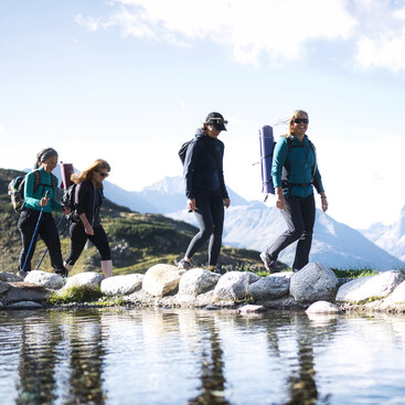 Vier Frauen wandern mit Rucksäcken und Isomatten entlang eines alpinen Wasserbeckens. Sie überqueren eine flache Steinbrücke bei Sonnenschein. Im Hintergrund erstrecken sich Bergketten.