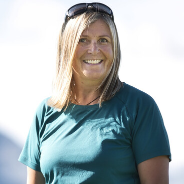 Geli Häusl mit schulterlangen blonden Haaren und Sonnenbrille am Kopf. Sie trägt ein petrolfarbenes Sportshirt. Der Hintergrund ist unscharf und hell.