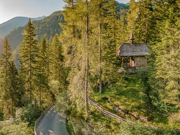 Kleine Kapelle aus Stein inmitten eines dichten Waldes in St. Anton, stimmungsvoll beleuchtet vom warmen Licht der tief stehenden Sonne.